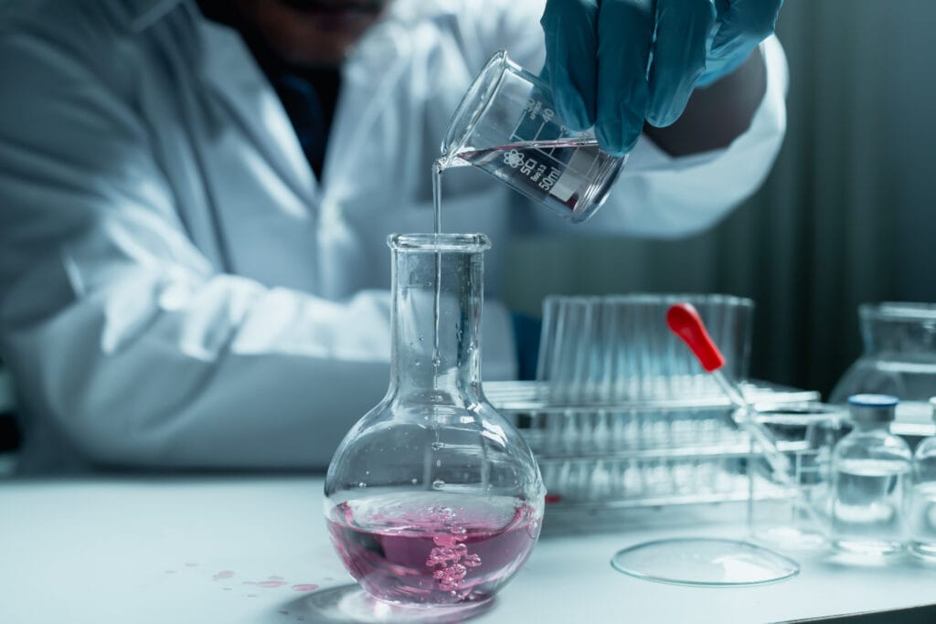 Scientist in a lab coat and gloves pouring a clear solution from a beaker into a round-bottom flask, with test tubes on the bench